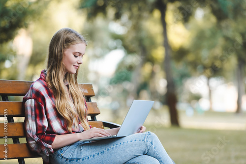selective focus of beautiful girl in casual clothes sitting on wooden bench in park and using laptop