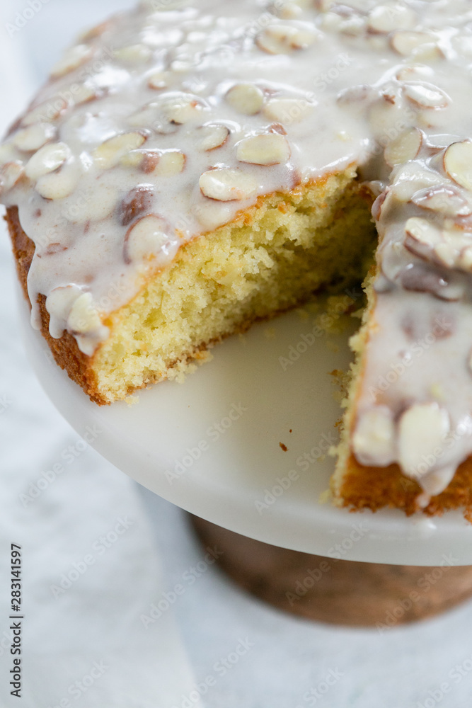 slice of almond olive oil cake with brown butter icing and sliced almonds, dripping icing on cake, marble and wood cake stand, styled bakery slice of almond olive oil cake with brown butter icing and sliced almonds, dripping icing on cake, marble and wood cake stand, styled bakery