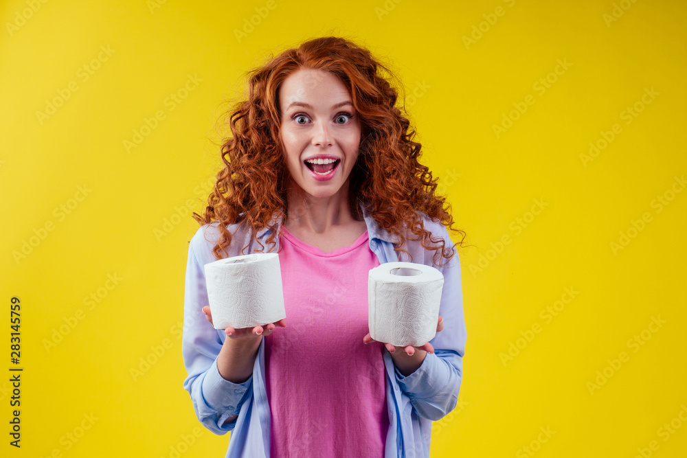 Beautiful curly redhead ginger woman choosing toilet paper roll on yellow color background Beautiful curly redhead ginger woman choosing toilet paper roll on yellow color background