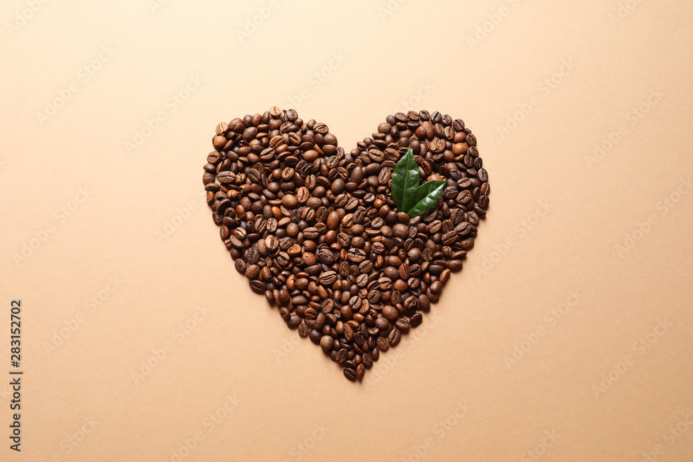 Heart shaped pile of coffee beans and fresh green leaves on light orange background, top view