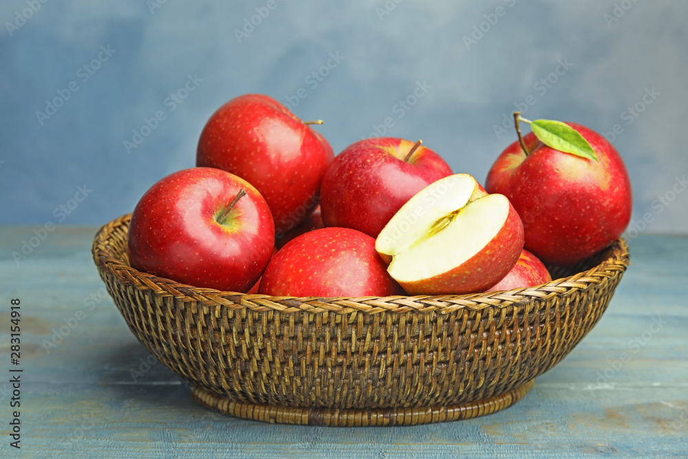 Wicker bowl with ripe juicy red apples on wooden table against blue background