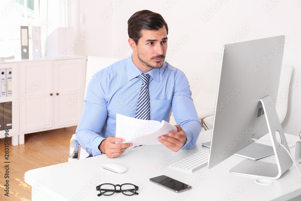 Handsome young man working with computer and papers at table in office Handsome young man working with computer and papers at table in office
