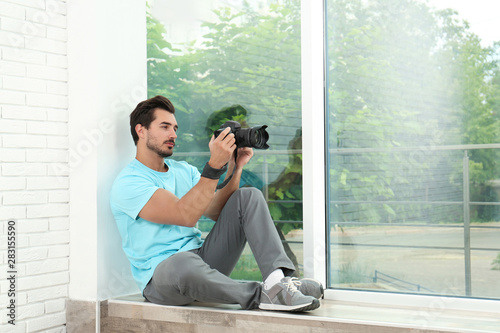 Young photographer with professional camera near window indoors. Space for text