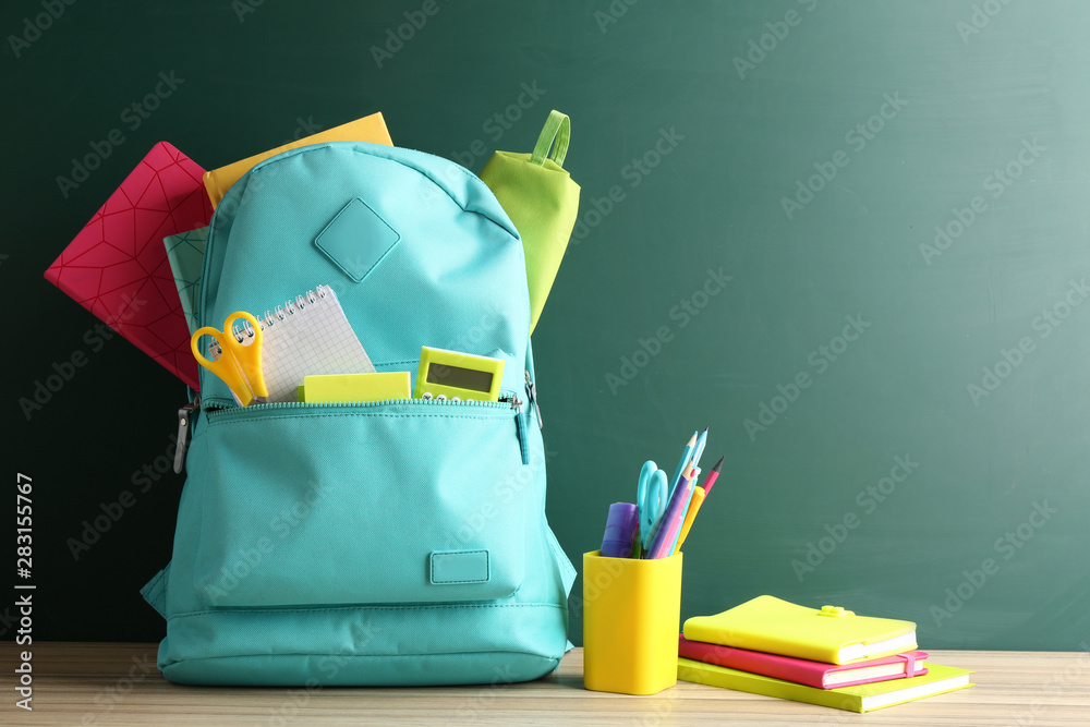 Backpack full of different school stationery on table near blank chalkboard. Space for text Backpack full of different school stationery on table near blank chalkboard. Space for text