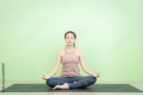 Beautiful smiling Asian woman sitting on black mat in lotus posture yoga beginning relaxation for meditation and exercise in the room with green background wall.