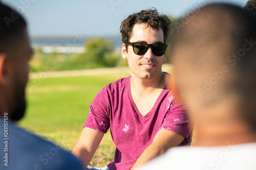 Smiling Caucasian man sitting on meadow with friends. Group of young people relaxing in park. Communication concept