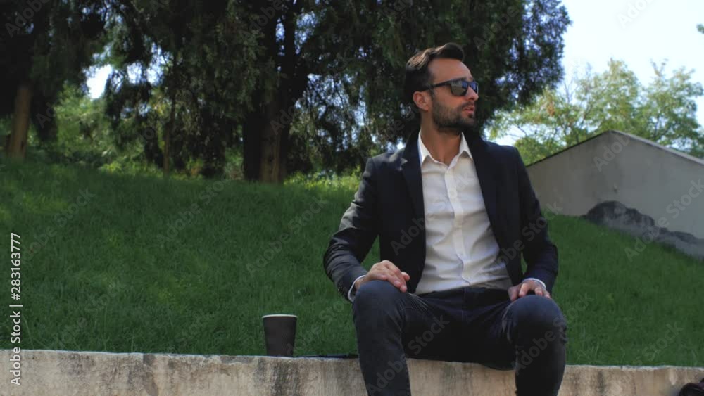 attractive man with a beard in a classic suit puts on sunglasses, then drinks tea or coffee from a disposable cup. 4k. 4k video