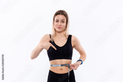 Young blond woman in a black sportswear with smart watches is happy pointing at the tape measure standing over white background. Checking waist after workout.