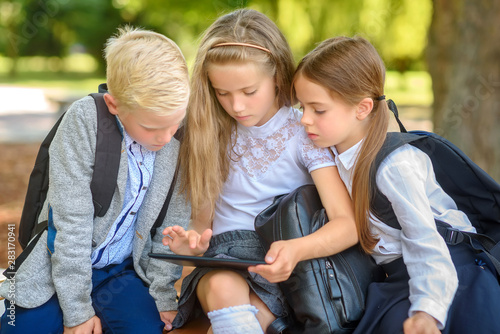 school friends, schoolchildren sitting on a bench in the park use a tablet to study lessons, social media communication