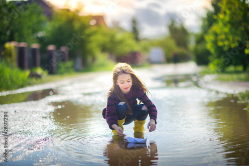 Fototapeta Naklejka Na Ścianę i Meble -  cute little girl in yellow boots plays in a puddle with a wooden boat, childhood, autumn concept