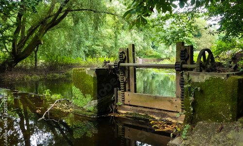 Landscape of a disused, Victorian sluice gate on the River Penk, Staffordshire. With rusty cogs and winding gear. Running water passing by, with overhanging branches and reflections on the surface. photo