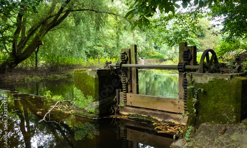 Landscape of a disused, Victorian sluice gate on the River Penk, Staffordshire. With rusty cogs and winding gear. Running water passing by, with overhanging branches and reflections on the surface.