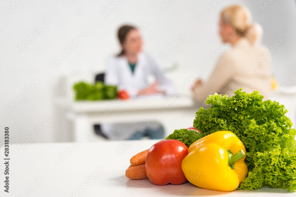 selective focus of dietitian in white coat and patient at table and fresh vegetables on foreground selective focus of dietitian in white coat and patient at table and fresh vegetables on foreground