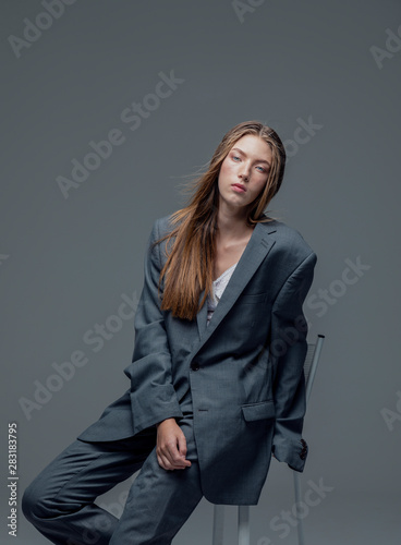 Portrait of weary young beautiful woman in oversized man's suit sitting isolated grey background