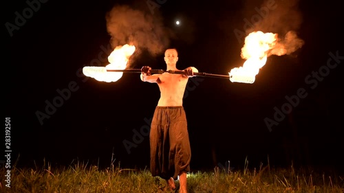 Low key. Young male with long hair and bare torso rotates burning torch outdoors on a black night video slow motion. Modern fakir does tricks with a burning staff