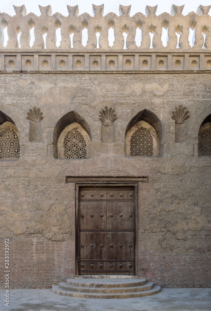 Wooden weathered door, perforated stucco window decorated with floral patterns, and three steps on stone bricks wall