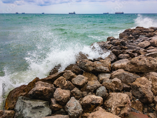 Sea waves with foam, beating large stones on the shore on a cloudy summer day with ships on the horizon.