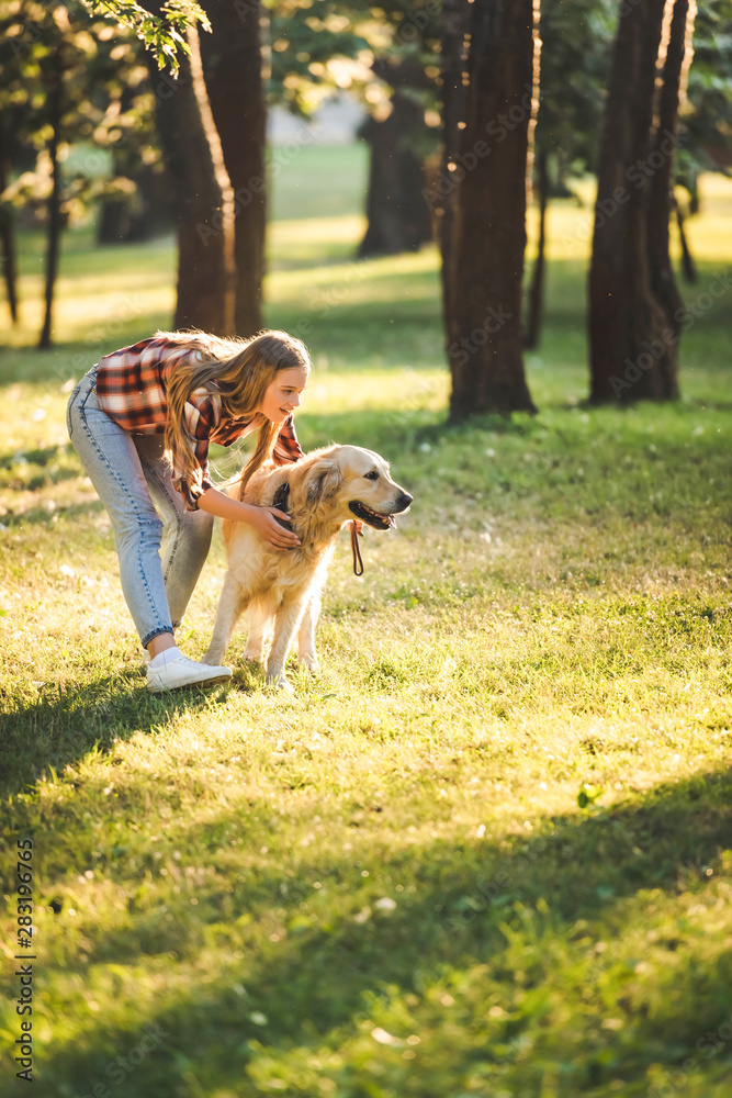 full length view of beautiful young girl in casual clothes hugging golden retriever while standing on meadow in sunlight