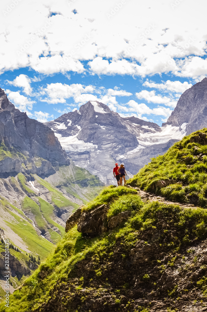 Couple walking on hiking path in the mountains captured on vertical photo. High snowcapped mountains in background. Summer adventure. Outdoor lifestyle concept. Trekking holidays