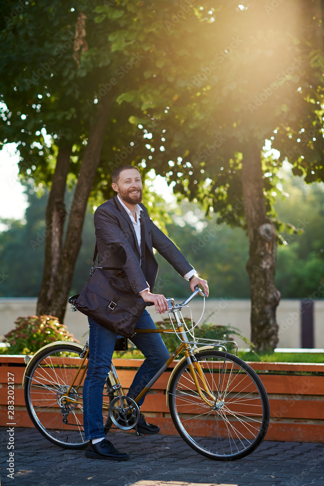 Obraz premium Bearded Businessman in business suit riding on retro bicycle to work on urban street in the morning on sunset