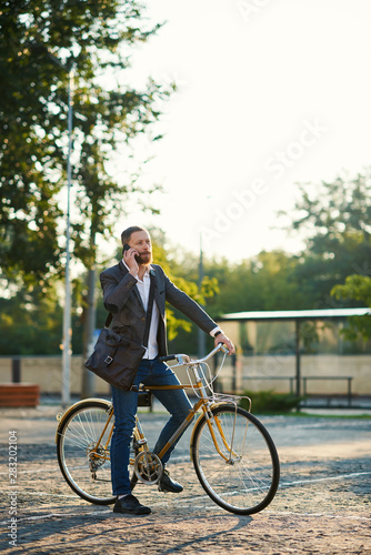 Bearded Businessman in business suit riding on retro bicycle to work on urban street in the morning on sunset. Drink coffee to go and speaking on mobile phone