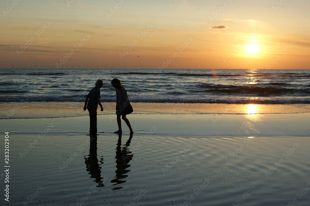 Silhouettes of two anonimous children against a sunset sky at the beach with reflection and windmills at the horizon