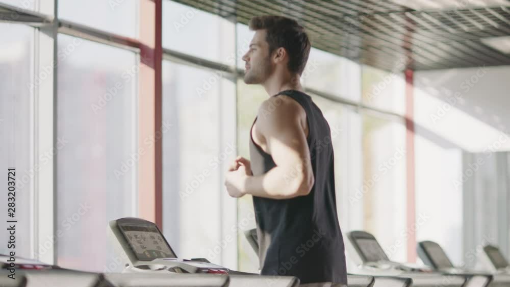 Cheerful man running on treadmill machine while cardio training in gym club.