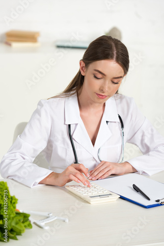 focused dietitian in white coat with stethoscope using calculator at workplace © LIGHTFIELD STUDIOS