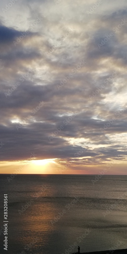 The autumn severe dark sea is illuminated by the setting sun, peeking through dense blue-gray clouds. Alarming and beautiful seascape. Dark gray clouds, solemn majestic sea and the golden setting sun.