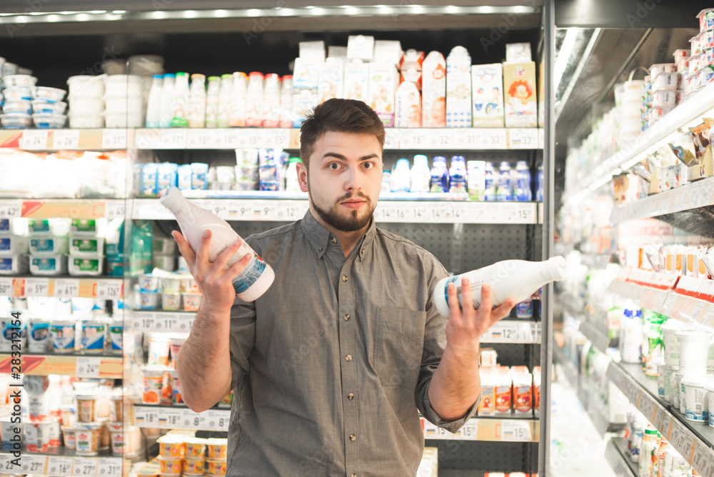 Man is standing with two bottles of milk in the milk department of the supermarket and can not choose. Lost man with a beard wearing a shirt chooses milk in a grocery store, looks at the camera.