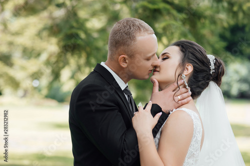 Stylish couple of happy newlyweds posing in the park on their wedding day. Perfect couple bride, groom posing and kissing. Young wedding couple enjoying romantic moments. © Tetiana Moish