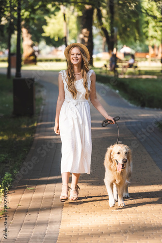 full length view of beautiful girl in white dress and straw hat walking with golden retriever on pathway and looking at camera