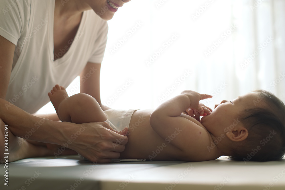 Obraz premium Asian young mother changing diaper for little baby lying on the soft mat at home. She looking and smiling to her kid with happiness while the child is playing and licking fingers. Family concept.