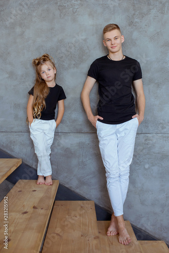 stylish caucasian siblings (brother and sister) in blak t-shirts and white pents standing by the gray wall in loft interior. Family, fashion, relatives, relationship and children concept. photo