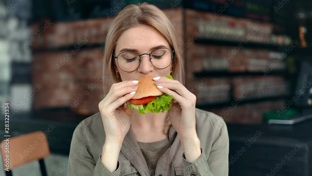 Pleasant woman biting desired fresh hamburger admiring amazing taste looking at camera Pleasant woman biting desired fresh hamburger admiring amazing taste looking at camera