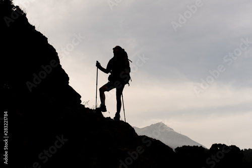 Silhouette slim girl standing on the rocks with hiking backpack and walking sticks