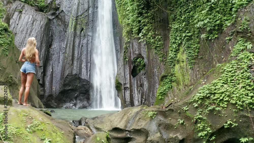Aerial drone view back of woman looking at the beautiful waterfall in green tropical rainforest in Bali, Indonesia Aerial drone view back of woman looking at the beautiful waterfall in green tropical rainforest in Bali, Indonesia