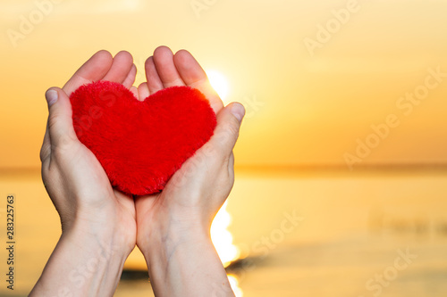 Woman's Hands holding Red fluffy plush Heart at the sunrise in front of the lake. Love, Romance and Friendship concept.