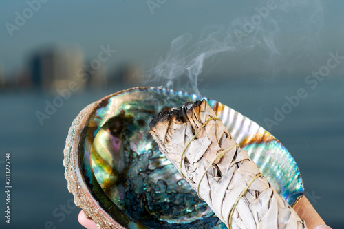 Smudging Ritual using burning thick leafy bundle of White Sage in bright polished Rainbow Abalone Shell on the beach at sunrise in front of the lake. photo