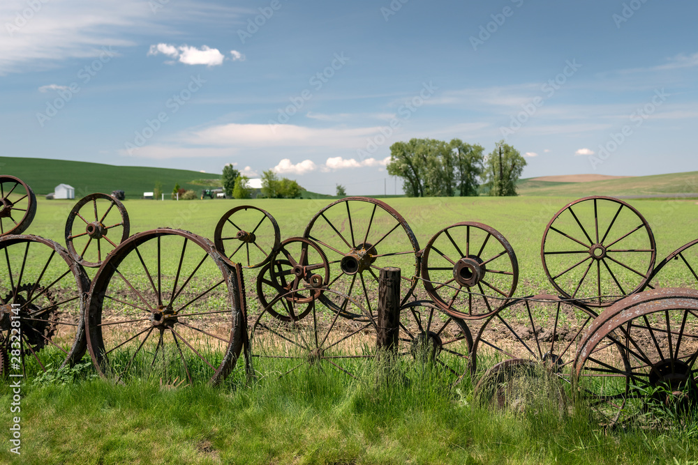 A fence made of of old metal rusted wheels. Green rolling hills, a farm, and a blue cloudy sky are in the background. Taken in the Palouse, Washington.