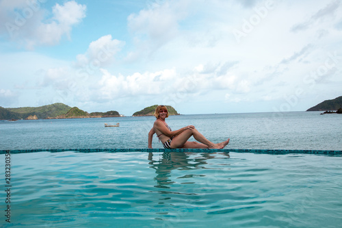 Smiling middle-aged woman in swimsuit sitting on the edge of the pool overlooking the turquoise sea
