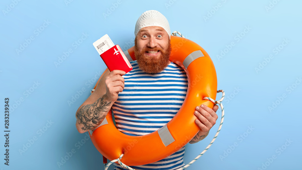 Cheerful bearded man holds passport with boarding pass tickets, has summer voyage, dressed in rubber bathingcap and sailor vest, feels excited about future trip, poses indoor. Traveling concept - obrazy, fototapety, plakaty 