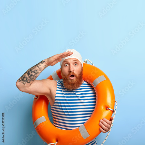 Emotional male lifeguard stares into distance, notices sinking person at sea, helps people to survive, carries inflated lifebuoy, stares with bugged eyes, wears rubber swimming cap, isolated on blue