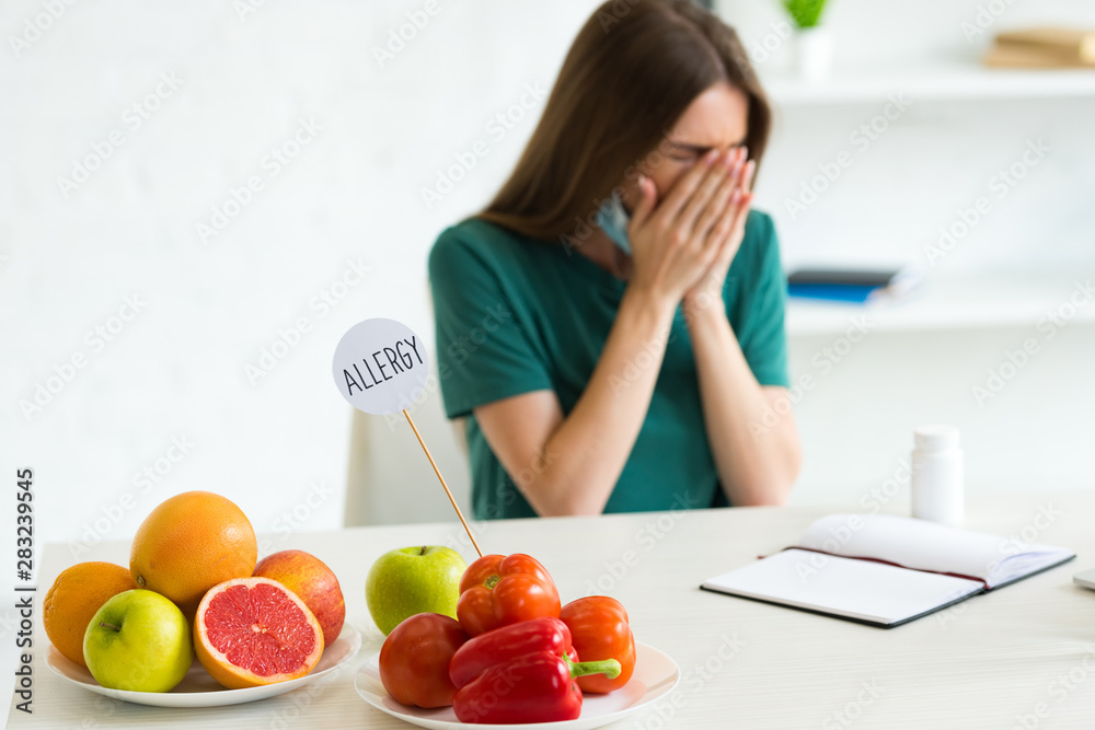 woman with medical mask sneezing while sitting at table with fruits, vegetables and pills