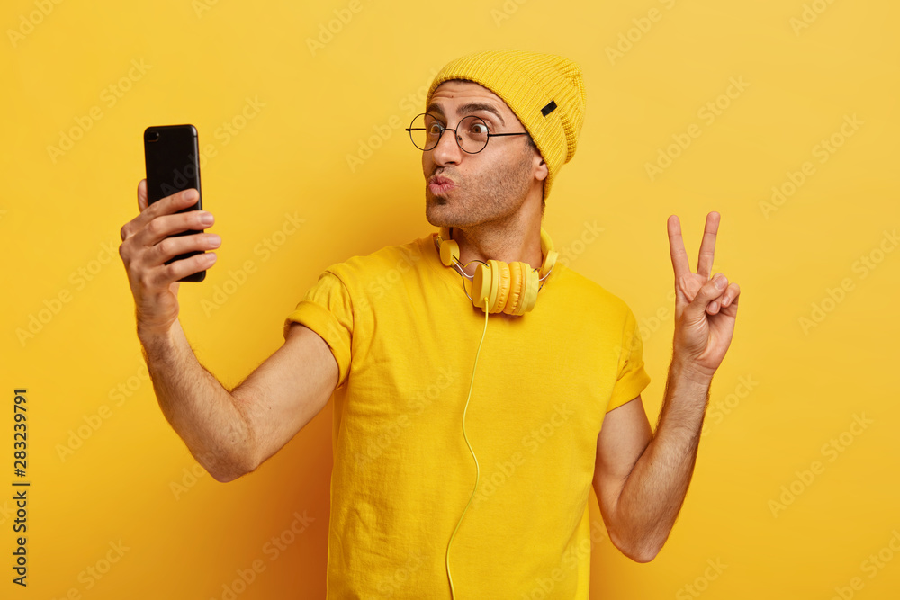 Handsome stylish hipster takes selfie, makes peace gesture, keeps lips folded, holds smart phone, wears transparent glasses, yellow hat and t shirt, stands indoor. Guy calls friend, poses at camera Handsome stylish hipster takes selfie, makes peace gesture, keeps lips folded, holds smart phone, wears transparent glasses, yellow hat and t shirt, stands indoor. Guy calls friend, poses at camera