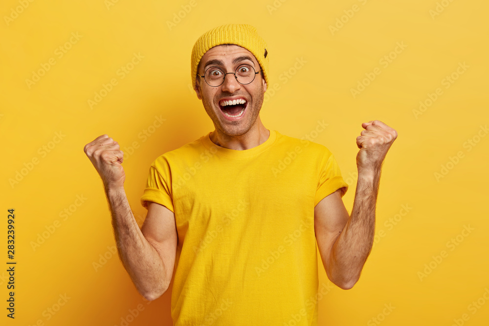 Hooray! Overemotive happy man makes fist pump from success and happiness, cheers achieving goal, wears optical glasses, fashionable yellow hat and tshirt, stands in studio, celebrates victory Hooray! Overemotive happy man makes fist pump from success and happiness, cheers achieving goal, wears optical glasses, fashionable yellow hat and tshirt, stands in studio, celebrates victory