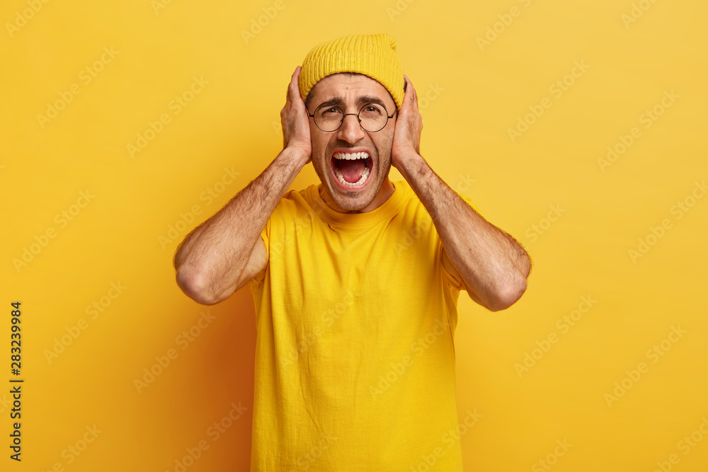 Depressed fed up stressful man looses temper, yells and screams loudly, covers ears, annoyed with loud unpleasant sound, being well dressed, isolated on yellow background, relieves from stress. Depressed fed up stressful man looses temper, yells and screams loudly, covers ears, annoyed with loud unpleasant sound, being well dressed, isolated on yellow background, relieves from stress.