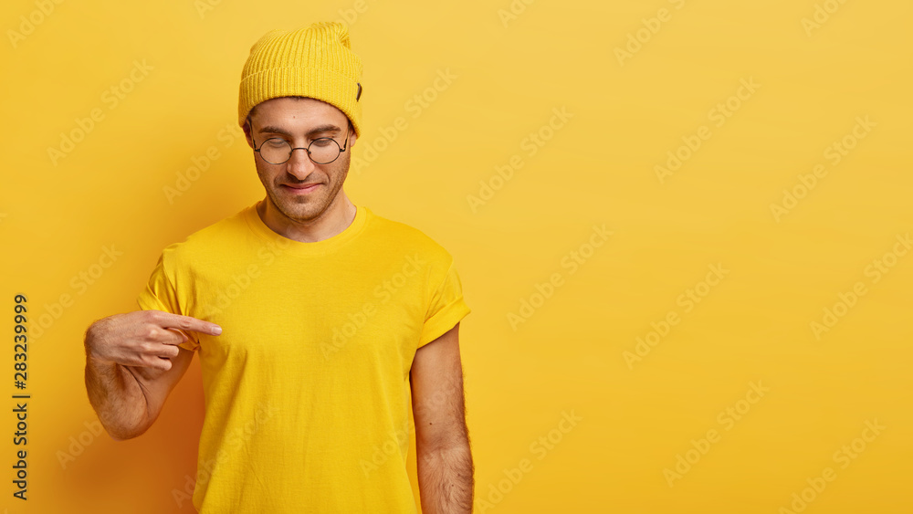 Satisfied male model points at blank space of his yellow t shirt, shows mockup place for slogan, advertisement or brand label, focused down, advertises clothes, likes yellow color, isolated.