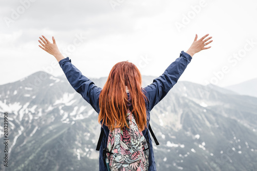 Successful young woman backpacker open arms on mountain peak. Pretty redheaded girl stands on spring snow mountains. Trekking and tourism concept. Copy space