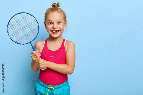 Indoor shot of glad red haired girl wears red vest and shorts, holds badminton racket, likes playing active games with friends, looks happily, isolated on blue background. Small child plays tennis © wayhome.studio 